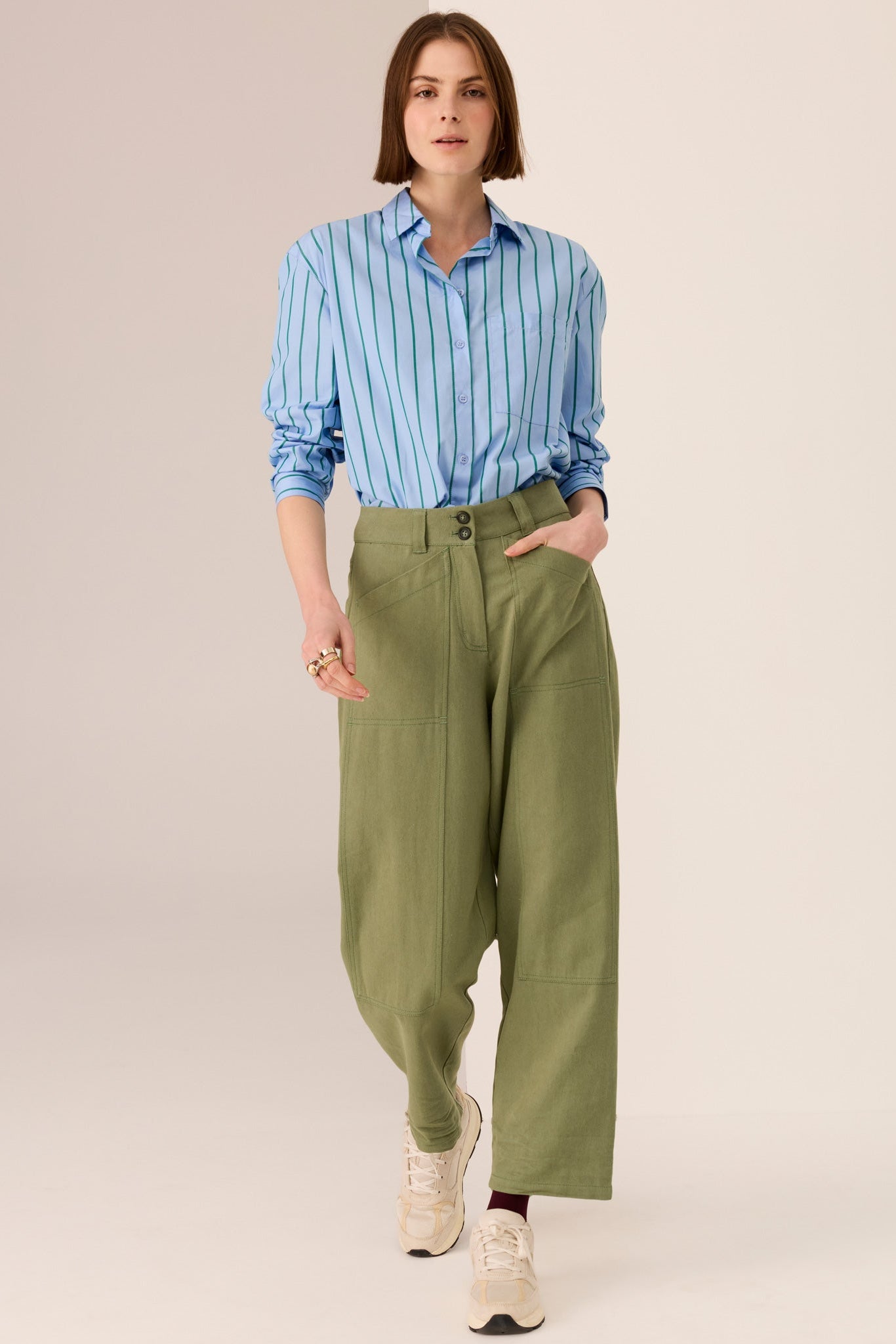 Woman wearing a blue shirt with green vertical stripe details and green pants on a white background. 