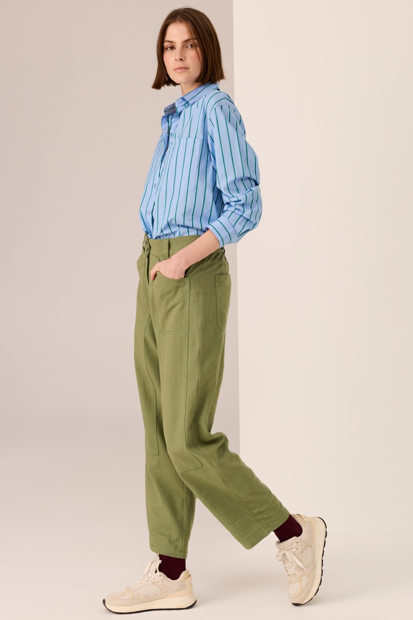 Woman wearing a blue shirt with green vertical stripe details and green pants on a white background. 
