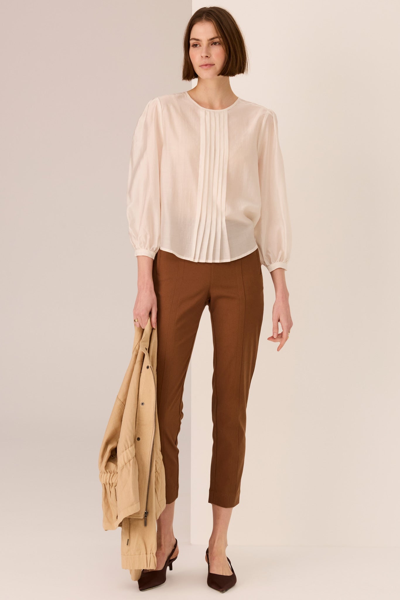 Woman wearing a ivory 3/4 sleeve blouse and brown pants holding a beige jacket on a plain background