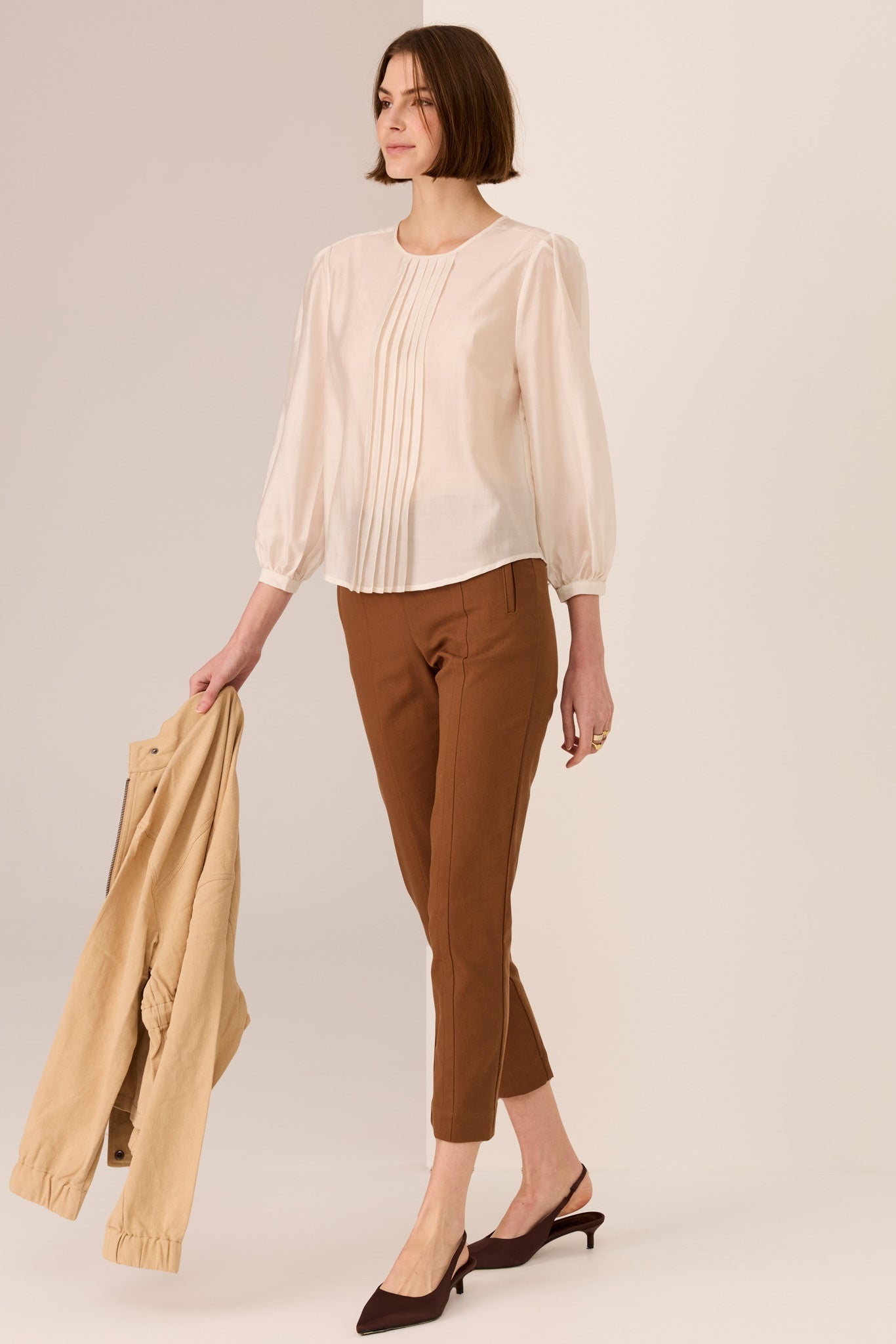 Woman wearing a ivory 3/4 sleeve blouse and brown pants holding a beige jacket on a plain background