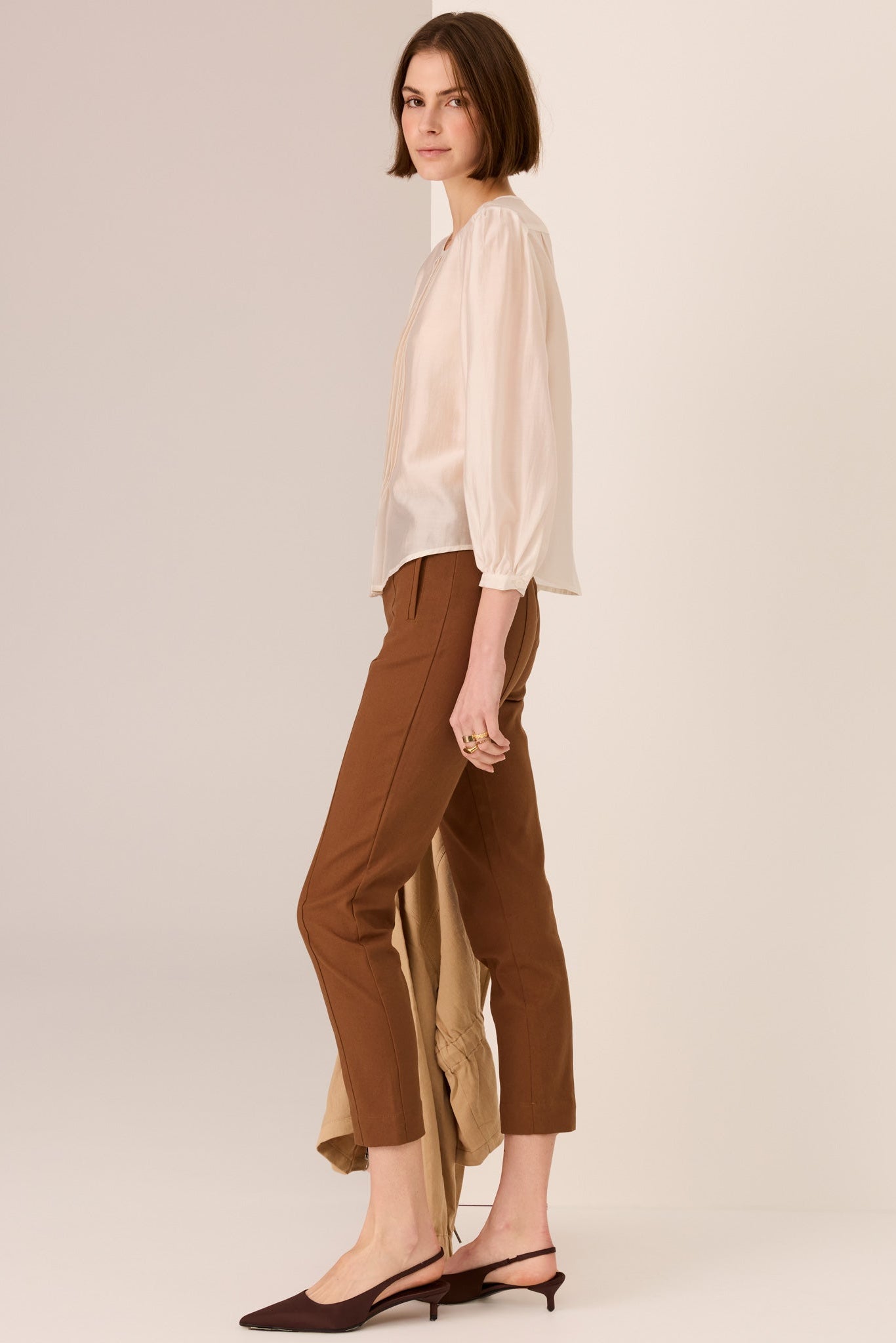 Woman wearing a ivory 3/4 sleeve  blouse and brown pants on a plain background