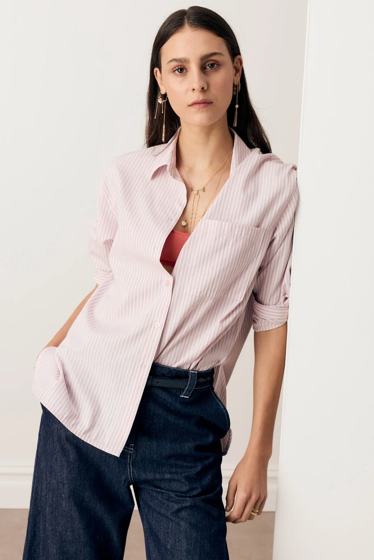 A woman wearing a light pink and white vertically striped button-down shirt over a rust-red top, paired with dark blue jeans, standing against a white wall.