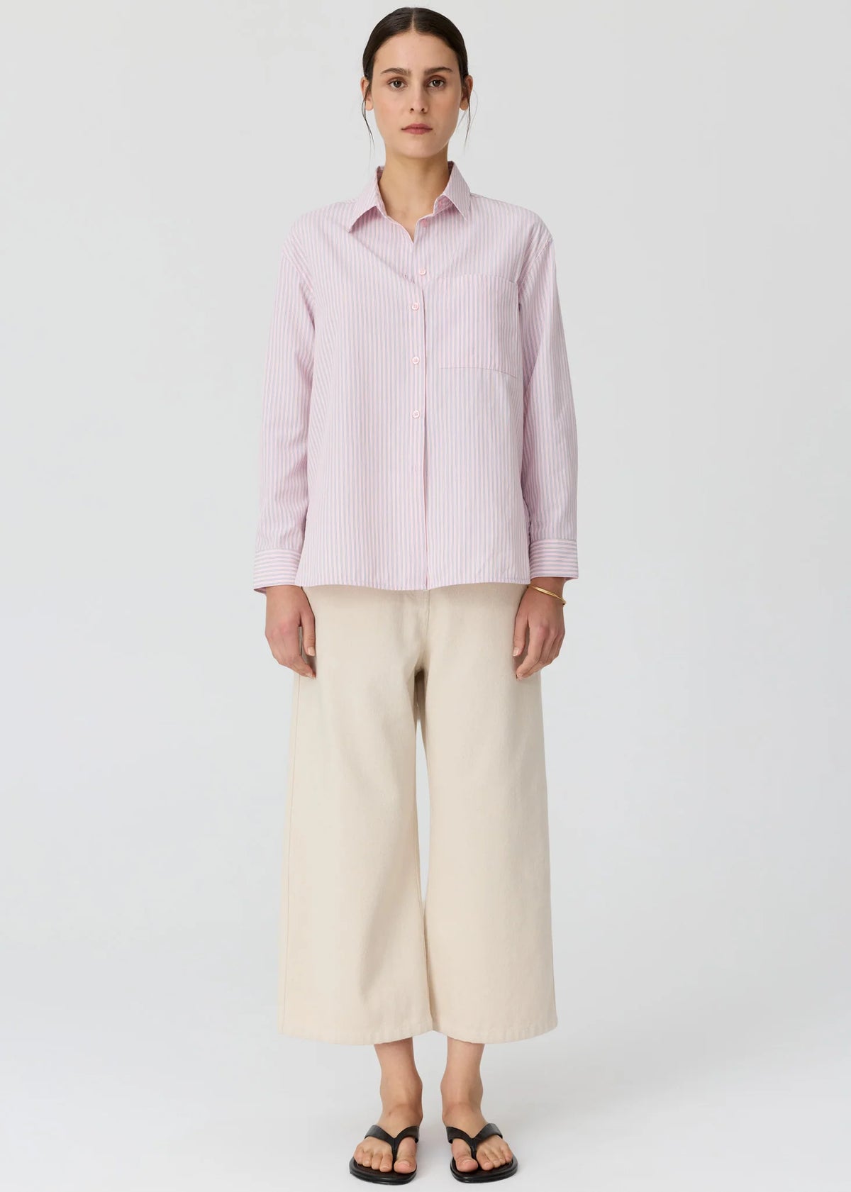 A model wearing a light pink and white vertically striped button-down shirt with cream wide-leg cropped pants and black flat sandals, set against a plain light gr3y studio background.
