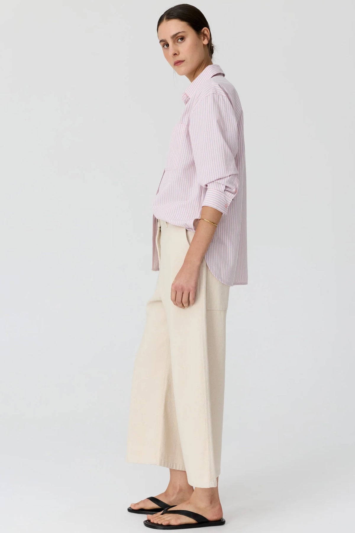 A model shown from the side wearing a light pink and white vertically striped button-down shirt with cream wide-leg cropped pants and black flat sandals, set against a plain light gr3y studio background.