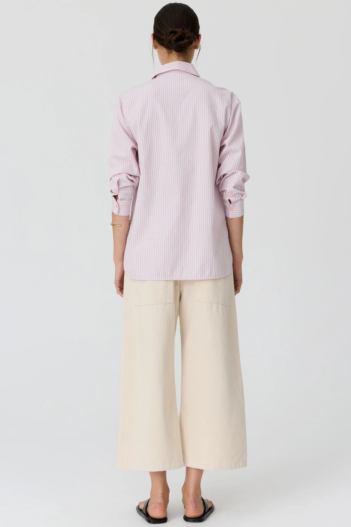 A model shown from the back wearing a light pink and white vertically striped button-down shirt with cream wide-leg cropped pants and black flat sandals, set against a plain light gr3y studio background.