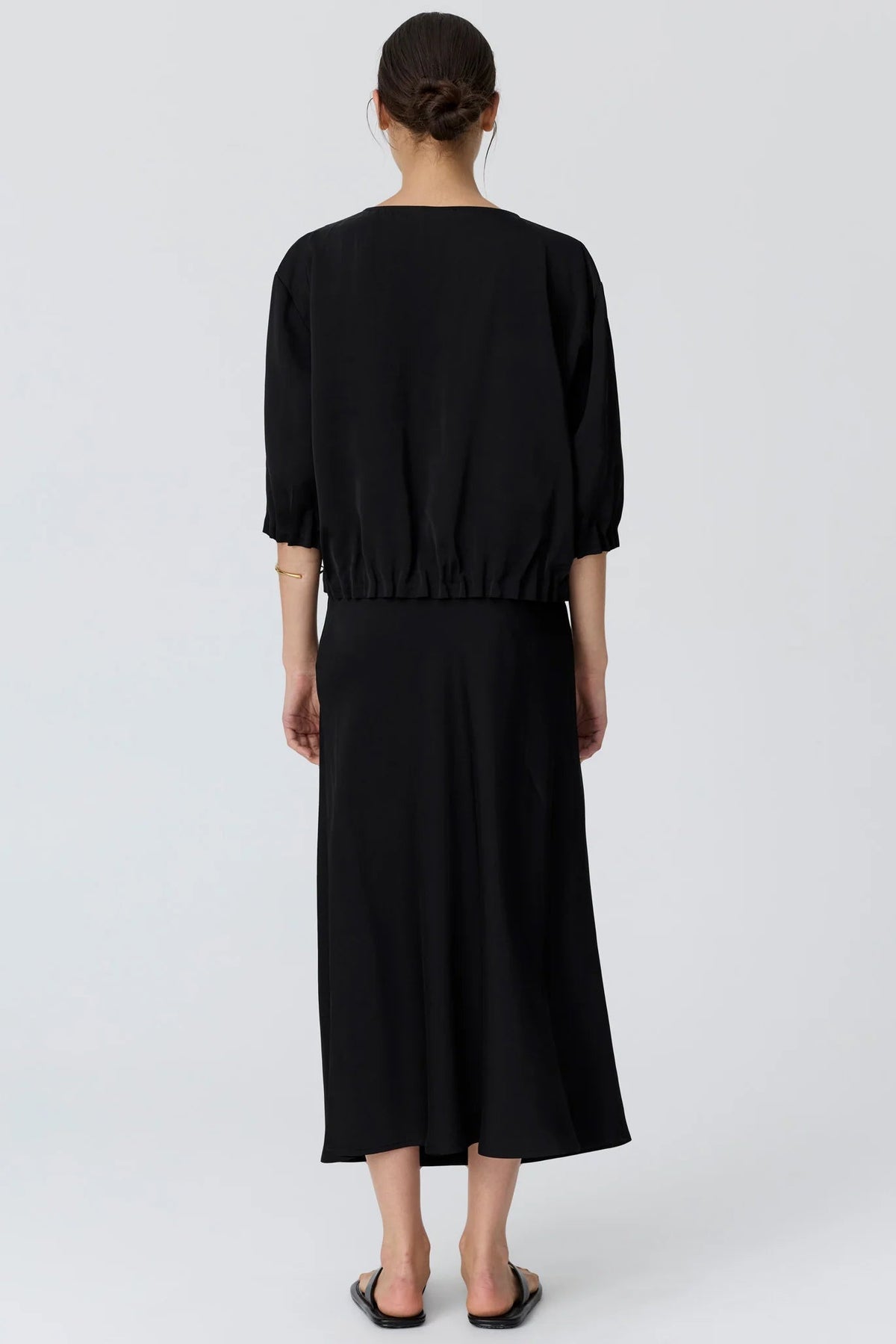 A woman shown from the back wearing a black top with voluminous elbow-length puff sleeves and matching skirt and black flat sandals, photographed against a light studio background.