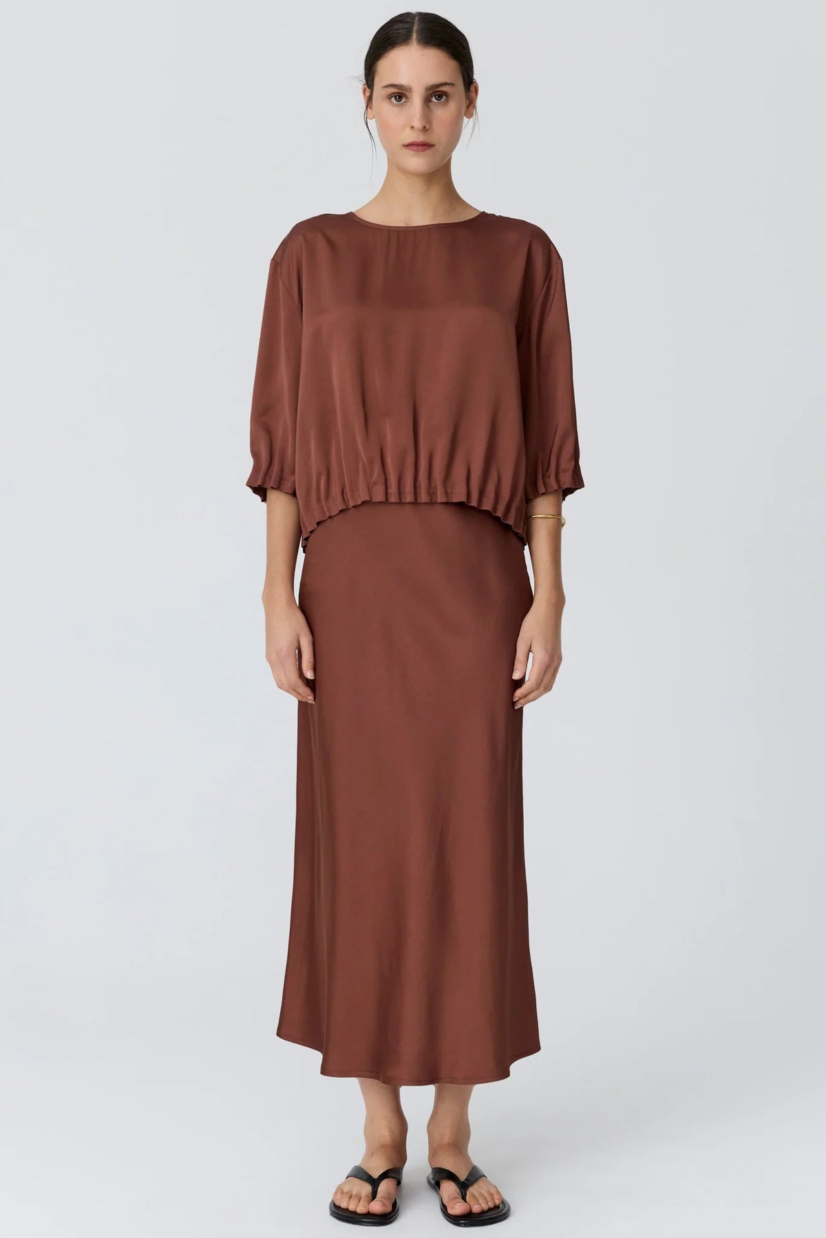 A woman modelling a toffee short puff-sleeve top and a matching midi skirt, paired with black flip-flop sandals, against a light studio background.