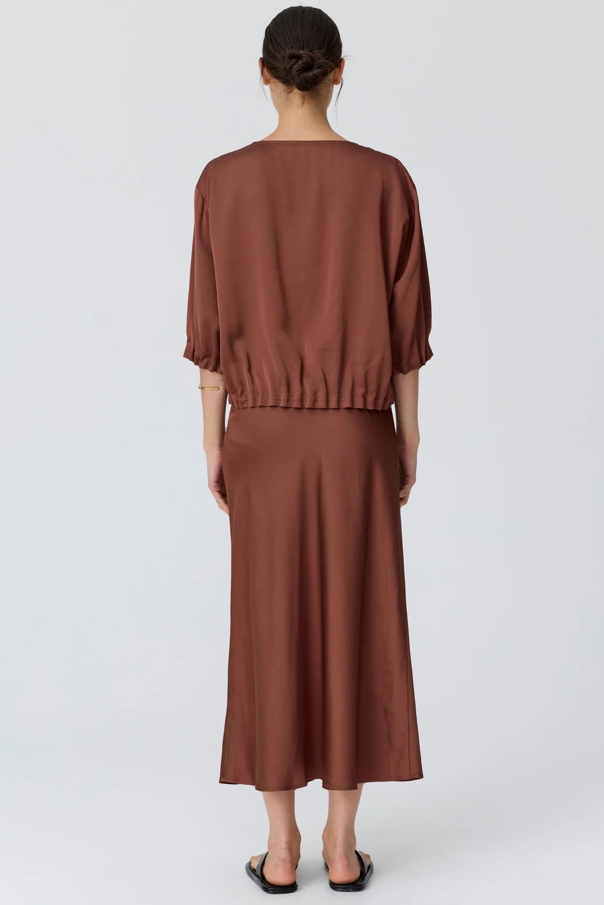 A woman in shown from the back wearing a toffee short puff-sleeve top and a matching midi skirt, paired with black flip-flop sandals, against a light studio background.