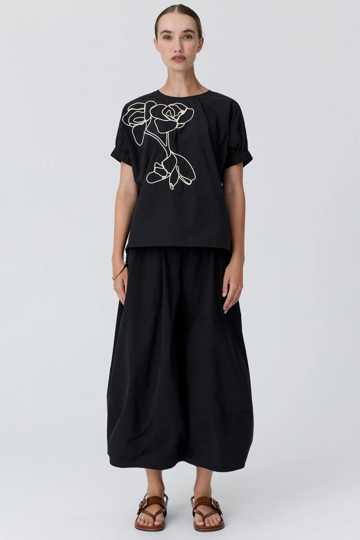 A woman modelling a black short-sleeve t-shirt with a white line-art rose graphic on the chest, paired with black bubble skirt and brown sandals, against a light studio background.