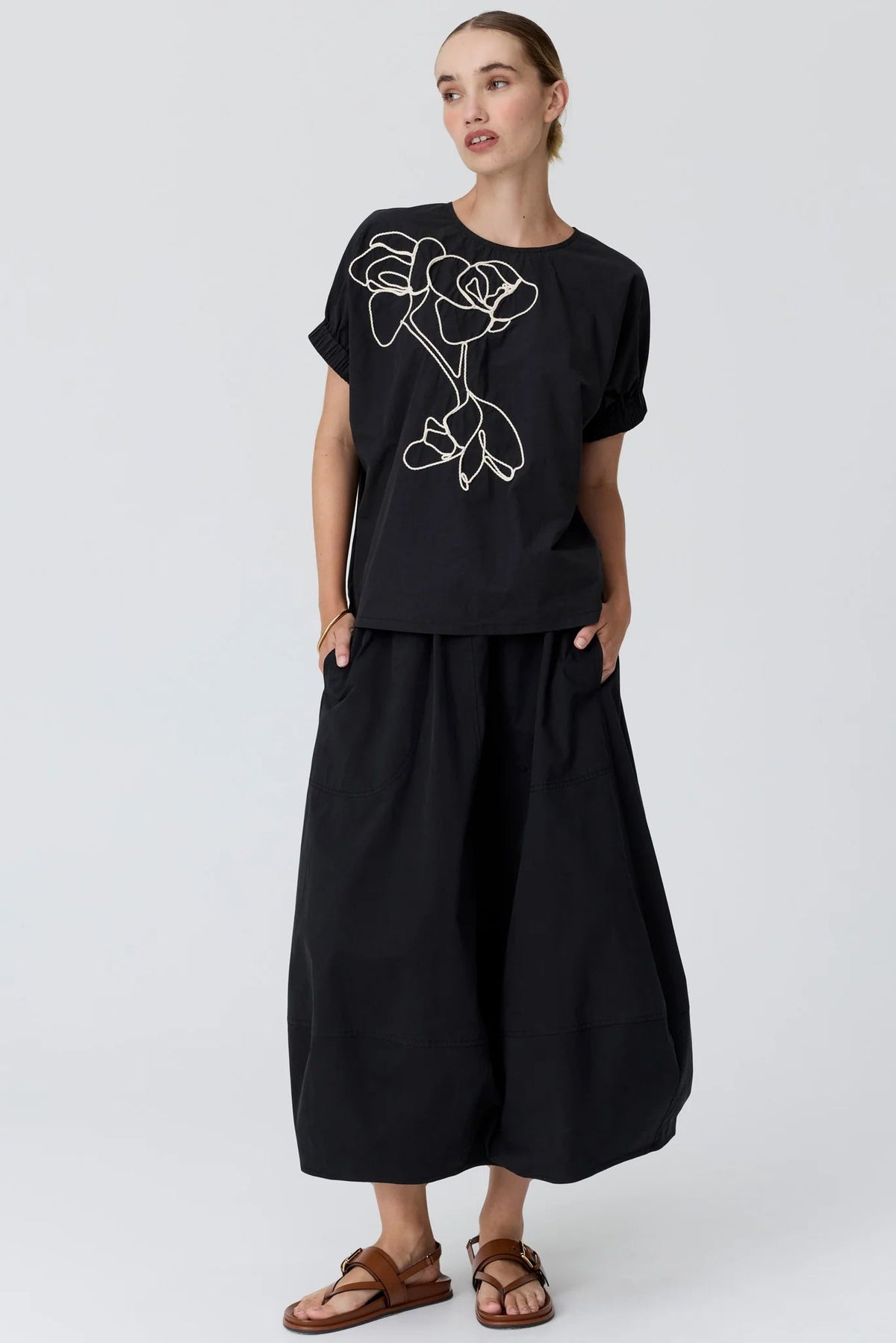 A woman modelling a black short-sleeve t-shirt with a white line-art rose graphic on the chest, paired with black Bubble skirt and brown leather sandals, against a light studio background.