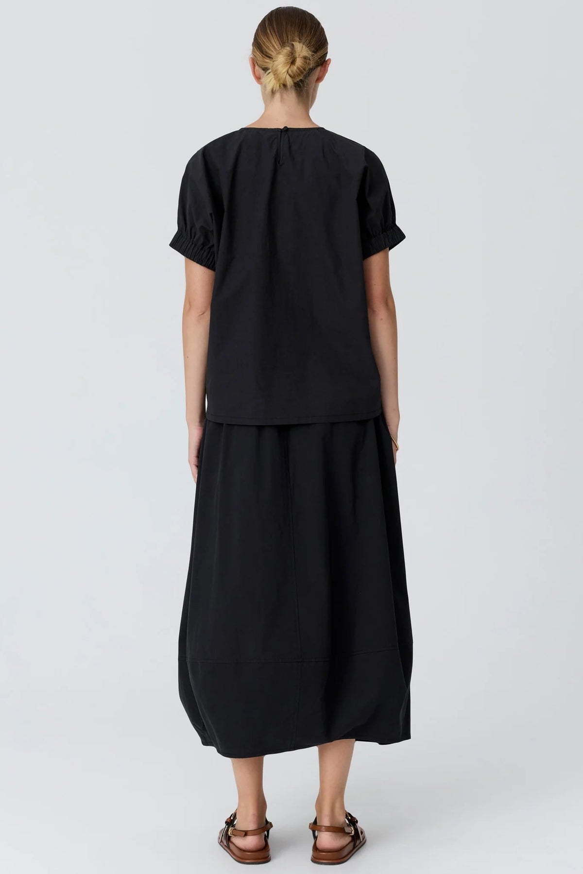 A woman viewed from the back wearing a black short-sleeve top and matching black midi skirt, paired with brown sandals, against a light gray studio background.