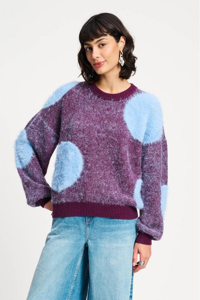 Person wearing a purple sweater with blue polka dots and light blue jeans on a white background