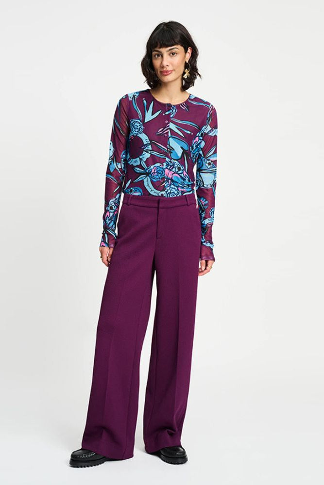 Woman wearing a patterned top and purple wide leg pants on a white background
