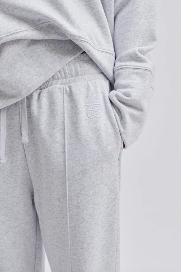 Model wearing a grey hoodie and grey track pant up close to show second female embroidery near left pocket. 