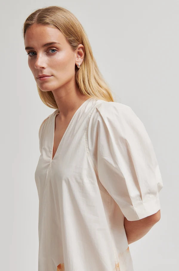 Model wearing a cream dress with puff sleeves and an abstract floral design on bottom left a plain background