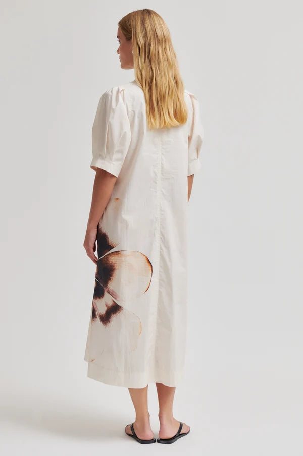 Model shown from the back wearing a cream dress with puff sleeves and an abstract floral design on bottom left a plain background