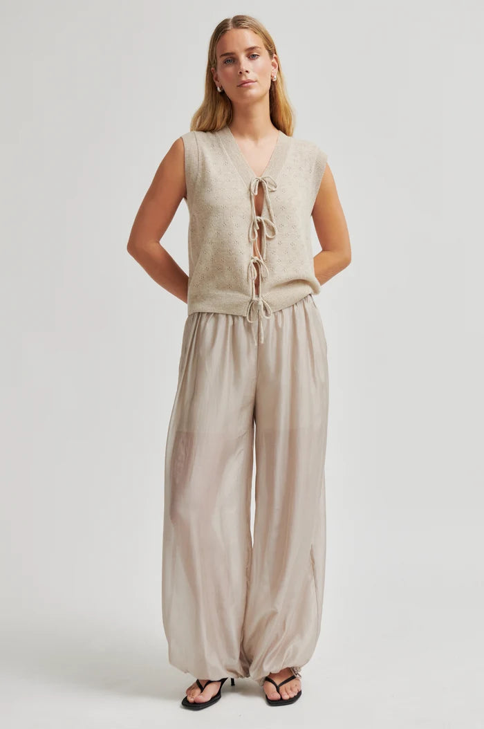 A woman modeling a sleeveless beige knit vest with front tie details and matching wide-leg beige pants, photographed against a plain light gray studio background.