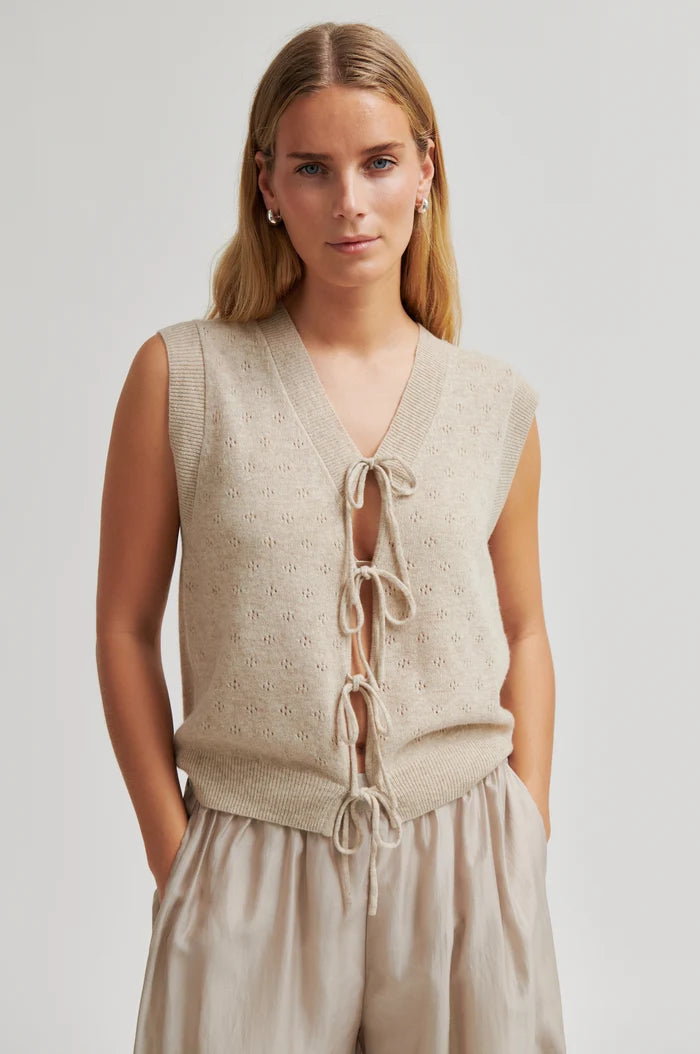 A woman wearing a sleeveless beige knit sleeveless vest with a V-neck and front tie closures, paired with matching beige bottoms, against a light grey studio background.