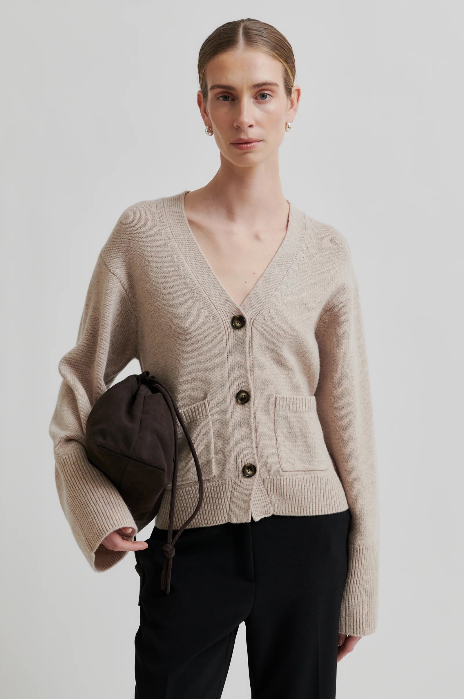 Woman wearing a beige cardigan holding a brown bag against a plain background