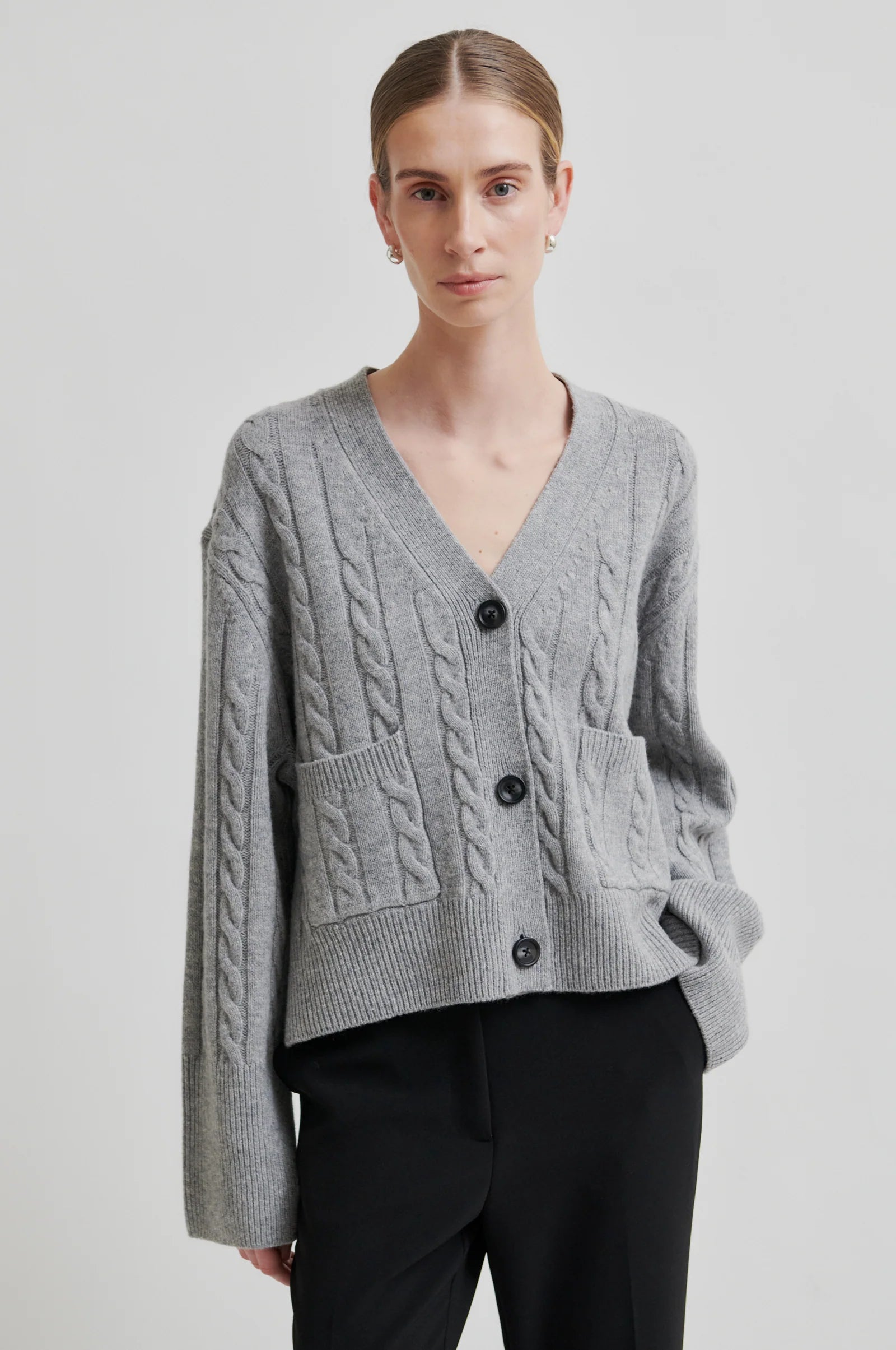 Model wearing grey long sleeve cable knit cardigan with black buttons and black pants on a grey background. 