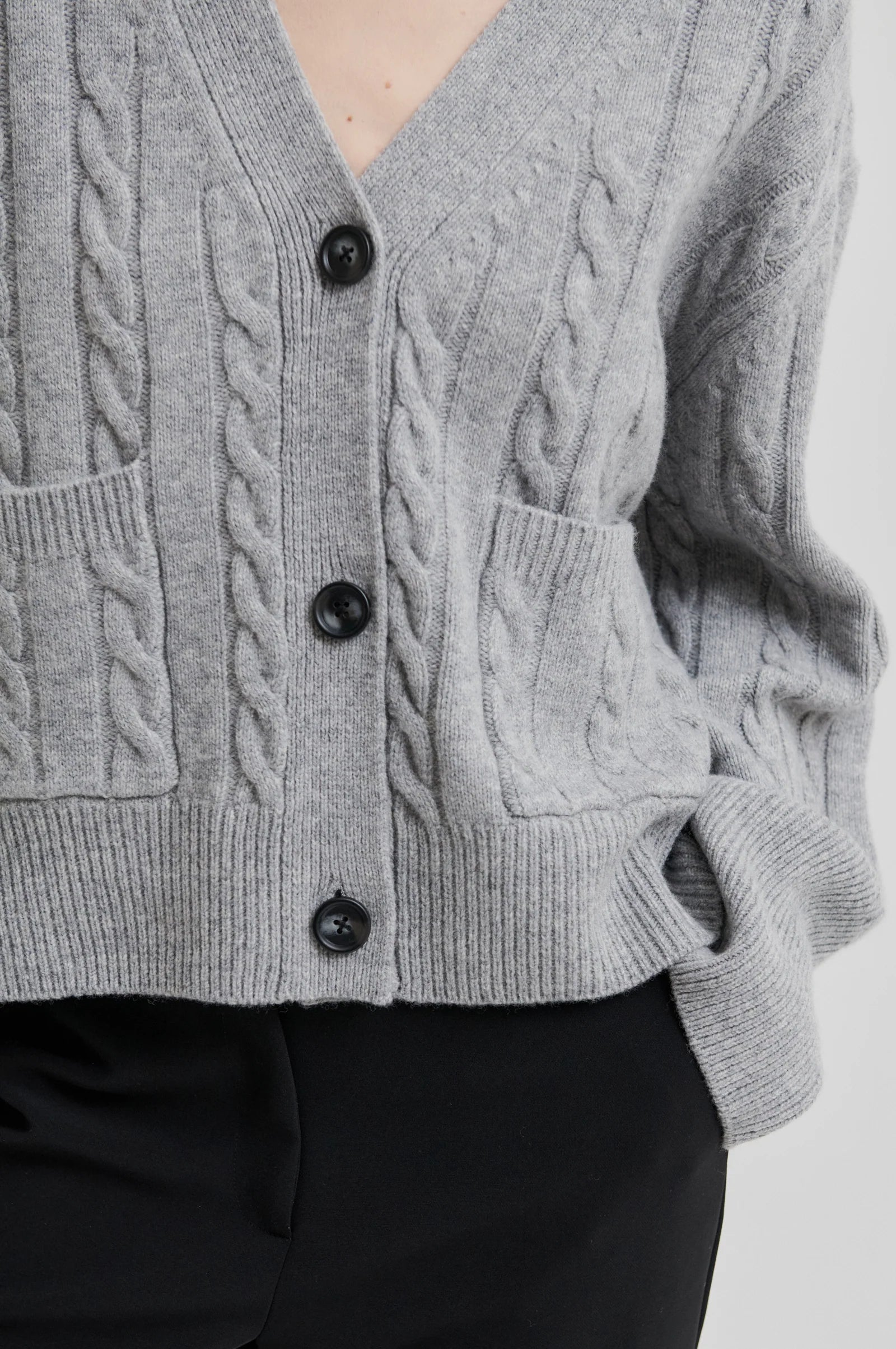 Model wearing grey long sleeve cable knit cardigan with black buttons and black pants on a grey background. 