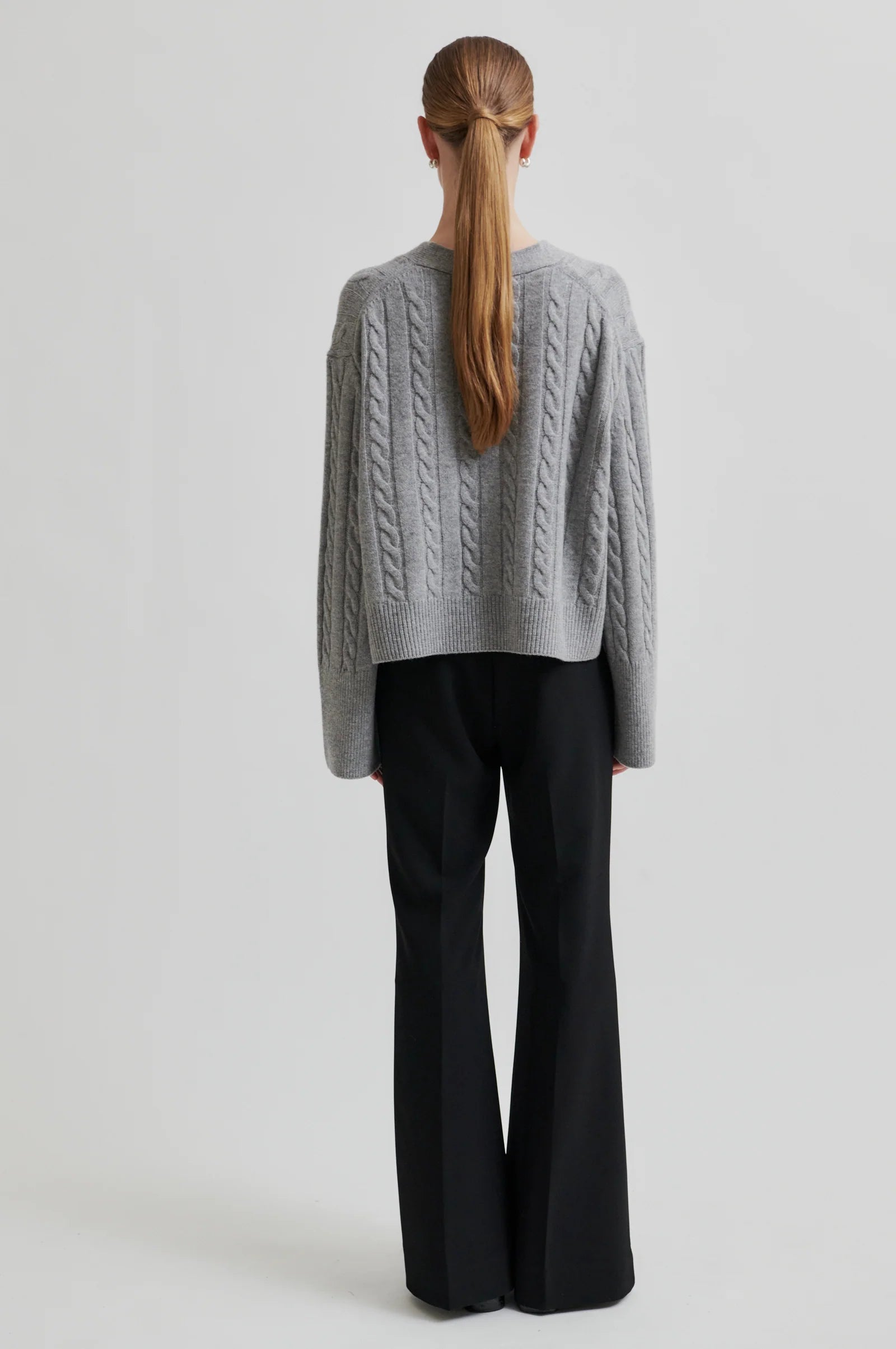 Model wearing grey long sleeve cable knit cardigan with black buttons and black pants on a grey background. 