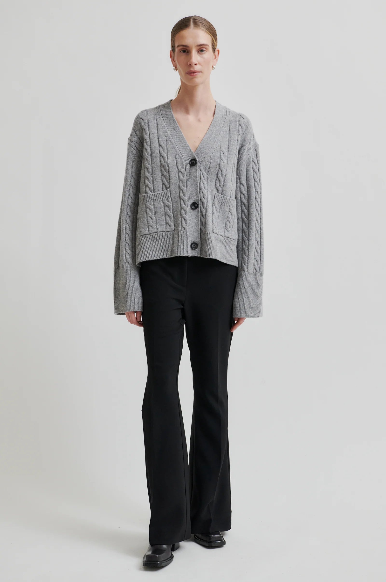 Model wearing grey long sleeve cable knit cardigan with black buttons and black pants on a grey background. 
