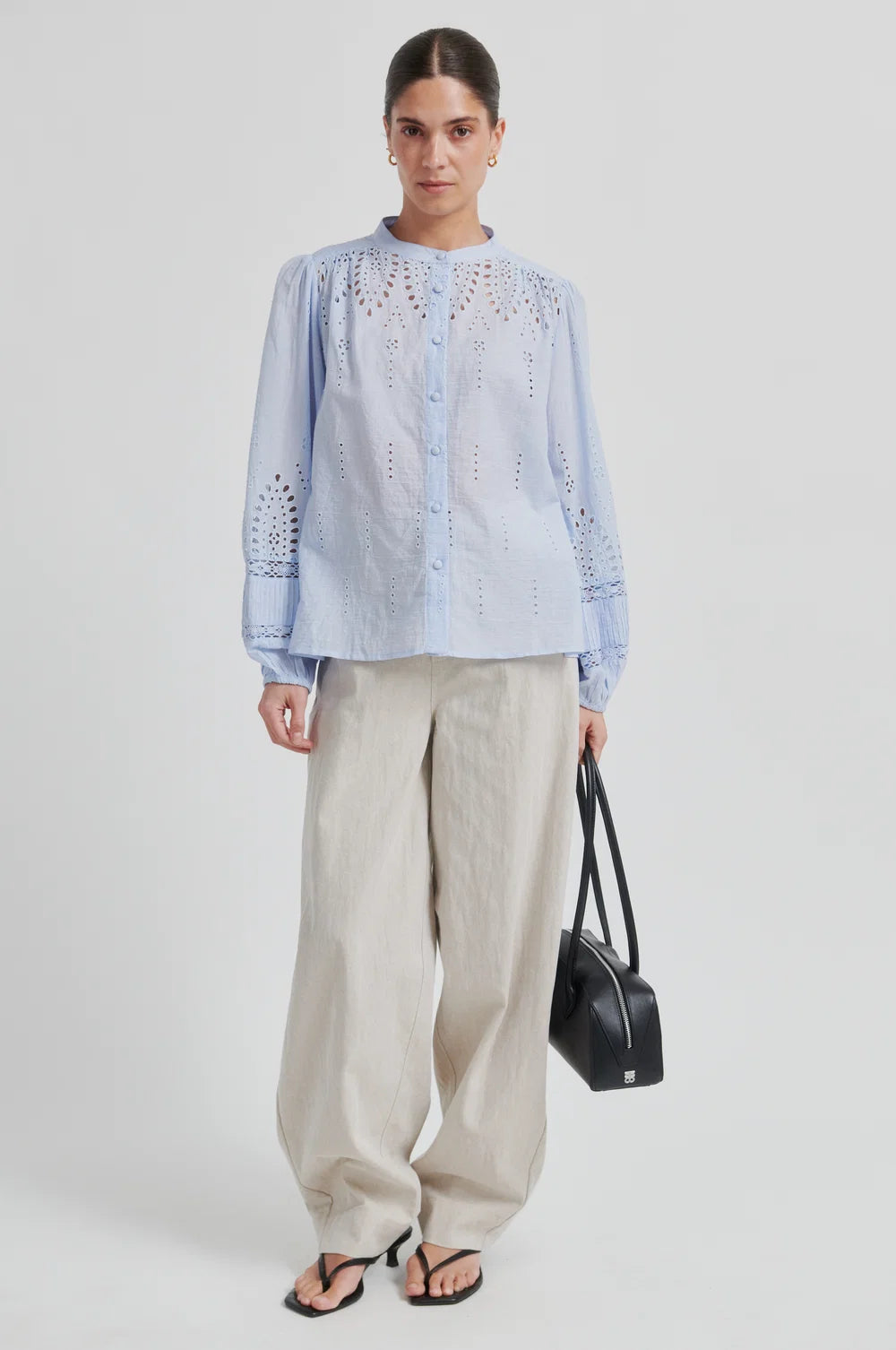 Woman  wearing a light blue blouse with intricate patterns and cream pants with a black bag and sandals on a plain background