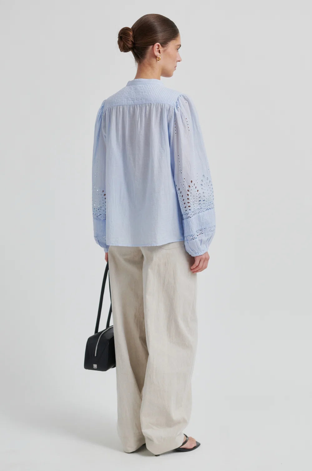 Woman from the back wearing a light blue blouse with intricate patterns and cream pants with a black bag and sandals on a plain background