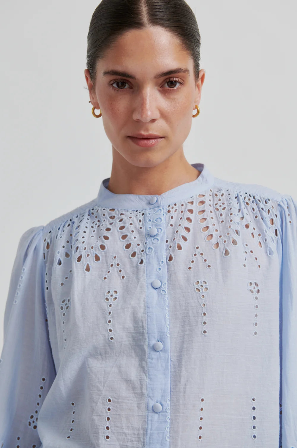 Woman up close wearing a light blue blouse with intricate patterns on a plain background
