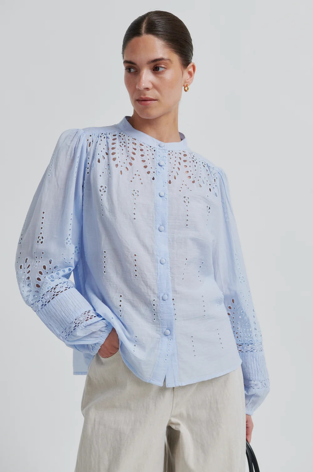 Woman wearing a light blue blouse with intricate patterns and cream pants on a plain background