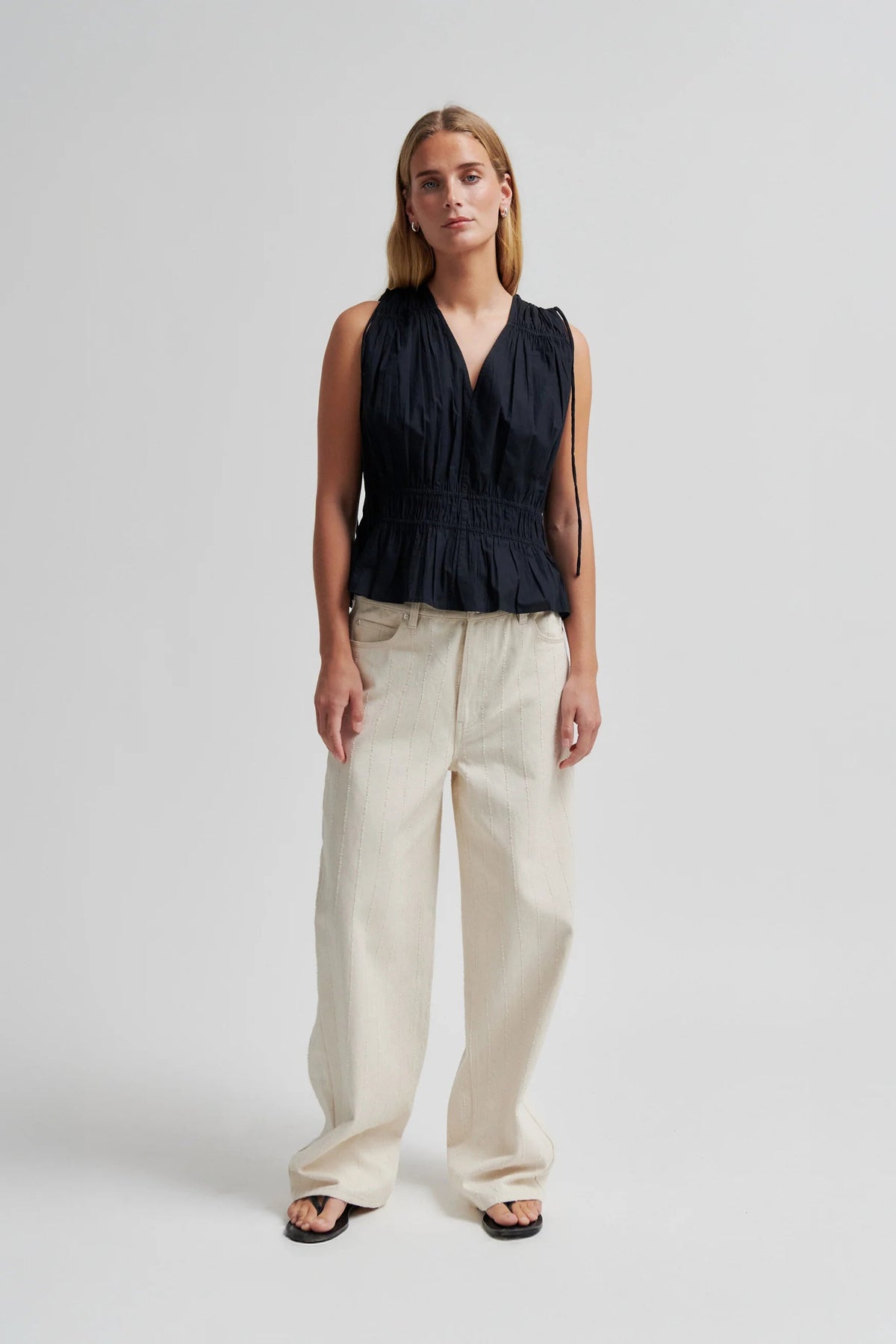 A woman stands against a light grey studio background wearing a sleeveless black top with a gathered waist and wide-leg cream pants.