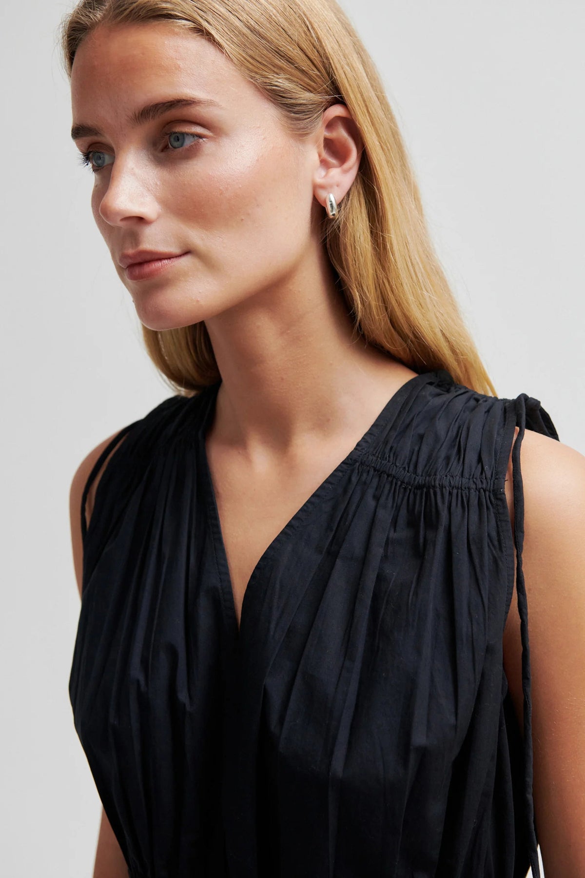 A close-up of a woman wearing a sleeveless black top with a gathered, pleated texture and a deep V-neckline, set against a plain light gray background.