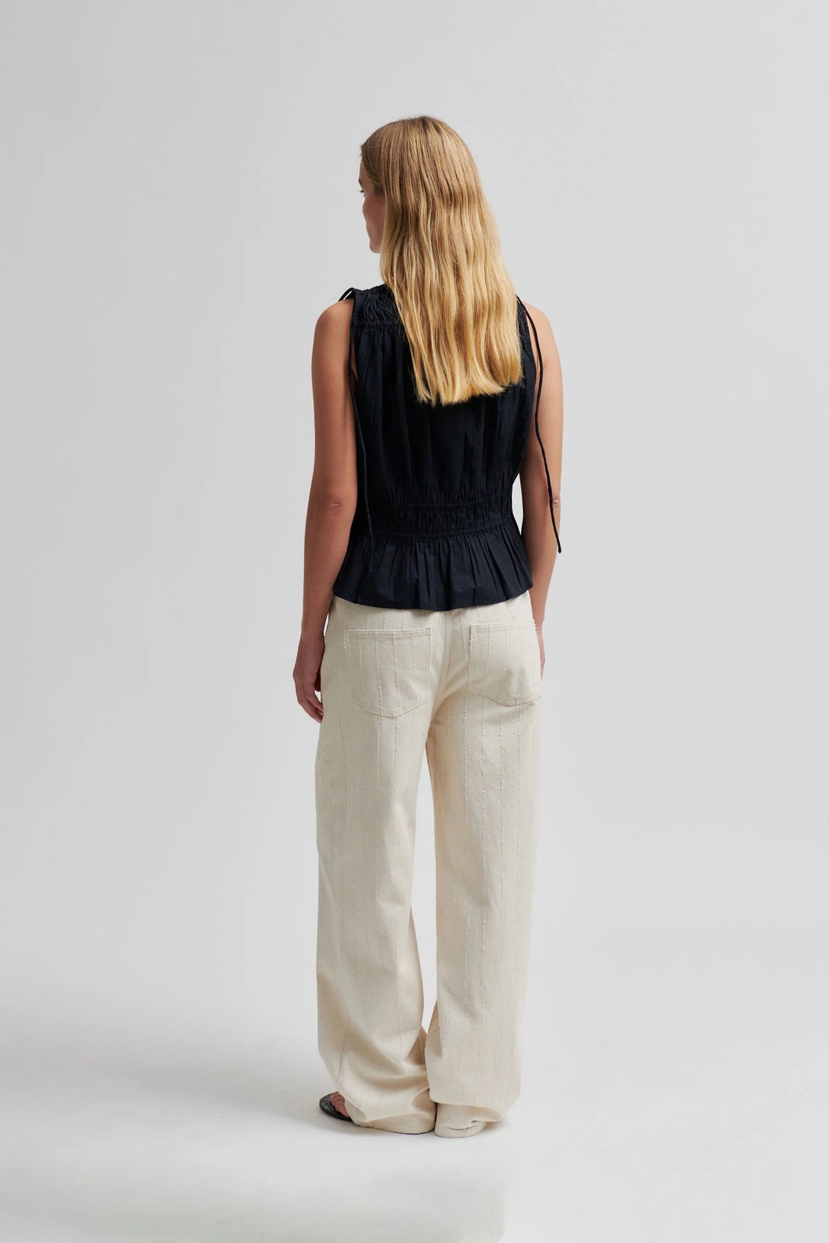 A woman stands with her back to the camera wearing a sleeveless black peplum-style top and wide-leg cream trousers against a plain light gray studio background.