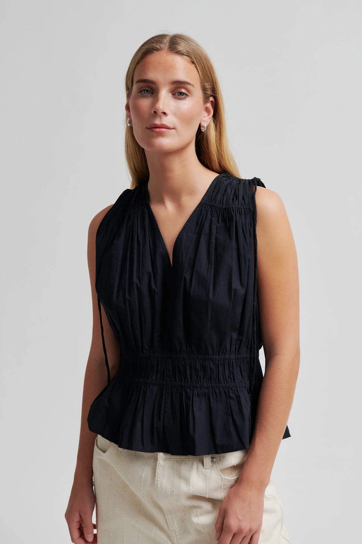 A woman modeling a sleeveless black peplum top with a gathered smocked bodice and V-neckline, paired with light beige trousers, against a solid light gray background.