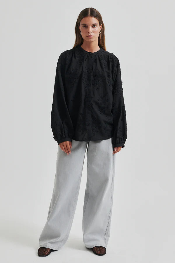 Model wearing a black oversized shirt with embroidery and light grey pants.