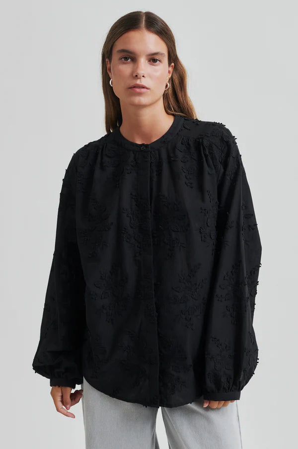 Model wearing a black oversized shirt with embroidery and light grey pants close up. 