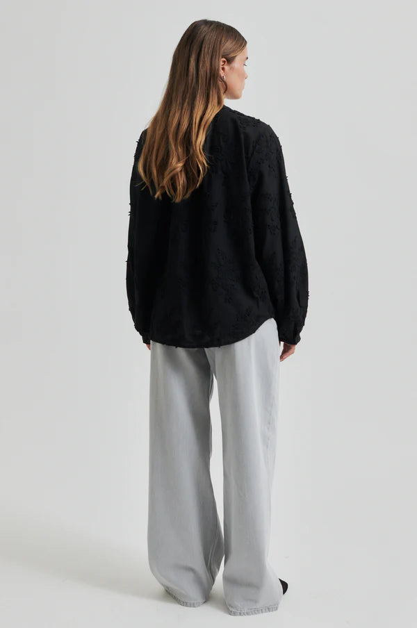 Model wearing a black oversized shirt with embroidery and light grey pants.