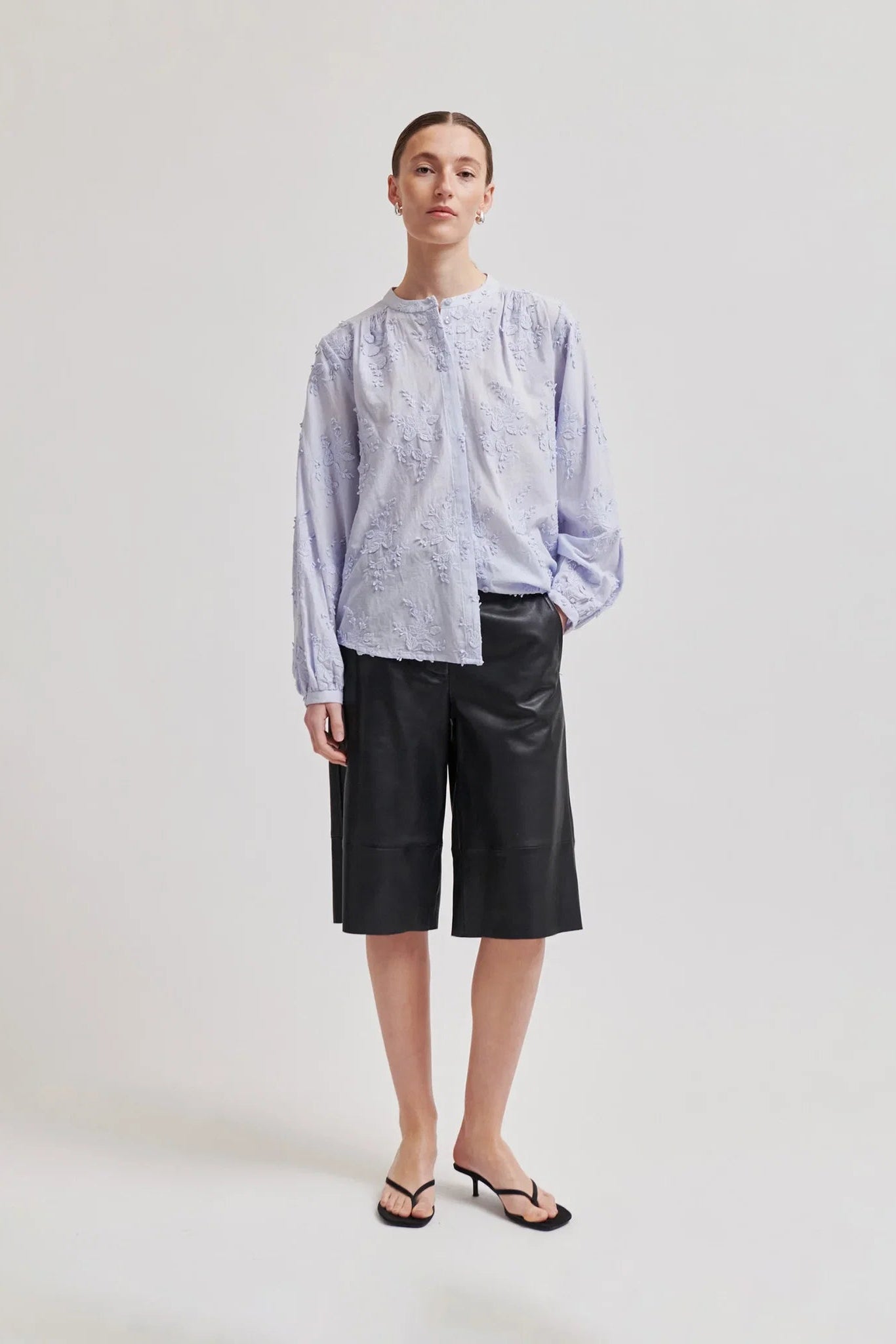 A model stands against a light gray studio background wearing a light blue and white embroidered long-sleeve blouse, black cropped leather-look shorts, and black heeled sandals.