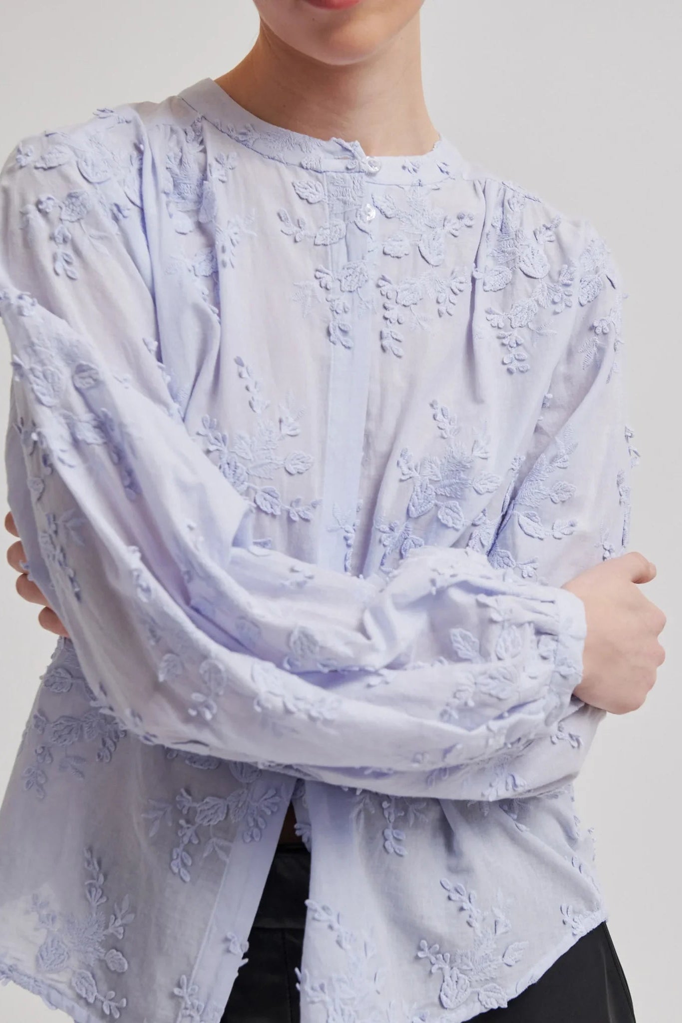 A close-up of a model wearing a light blue  long-sleeve blouse with intricate floral embroidery and puff sleeves, paired with black bottoms against a neutral light gray background.