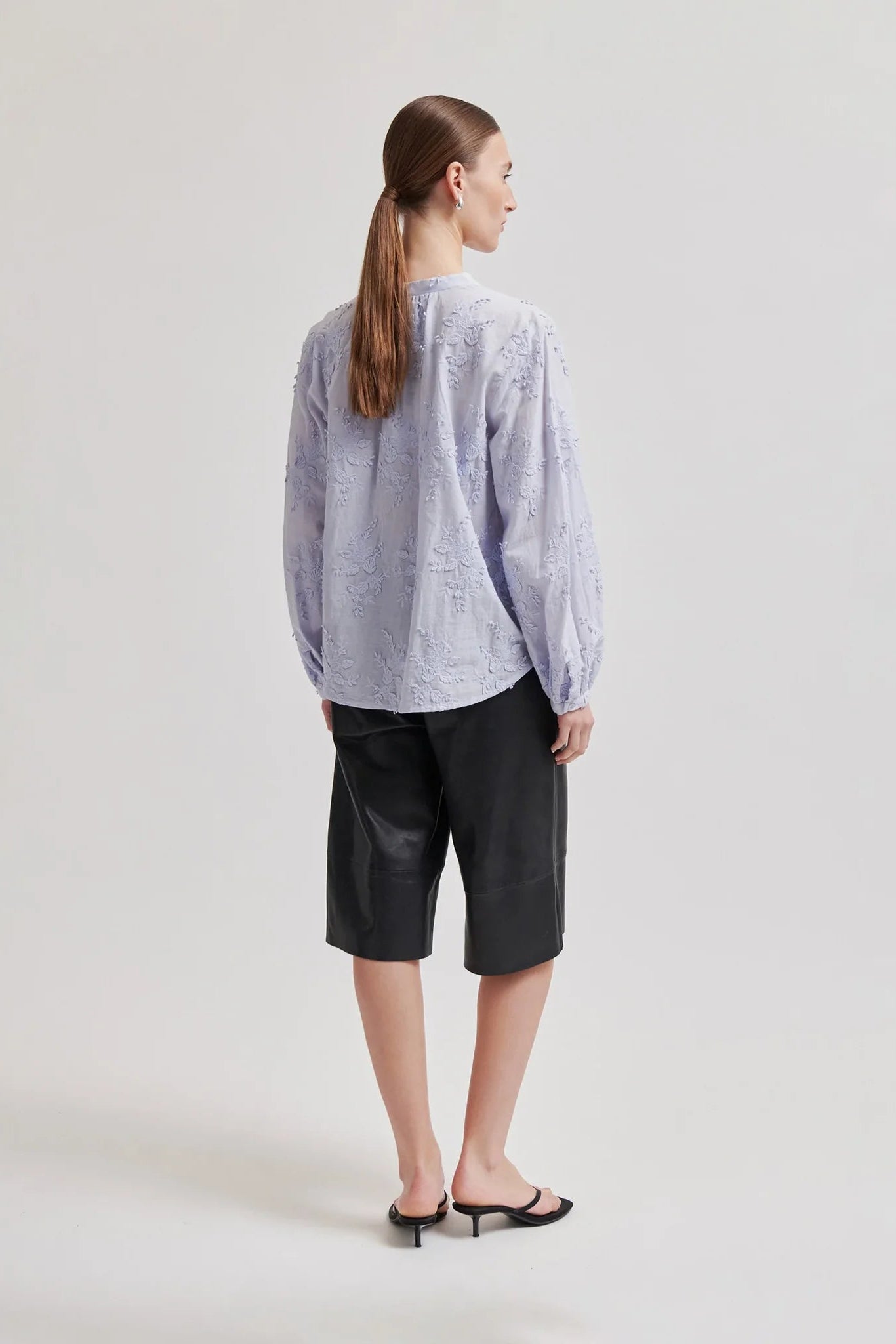 A woman is shown from the back wearing a light pale blue patterned long-sleeve blouse, black midi-length shorts, and black flat sandals against a plain light gray studio background.