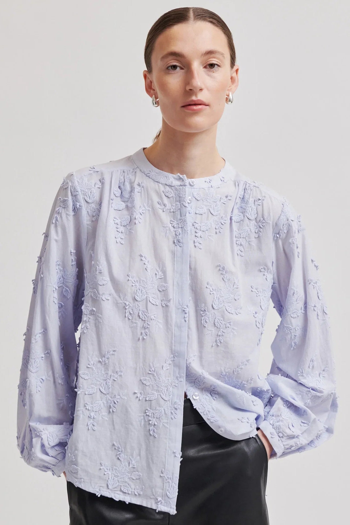 A model wearing a light blue long-sleeve blouse with textured floral embroidery and a button-up front, paired with black leather pants, against a neutral light gray background.