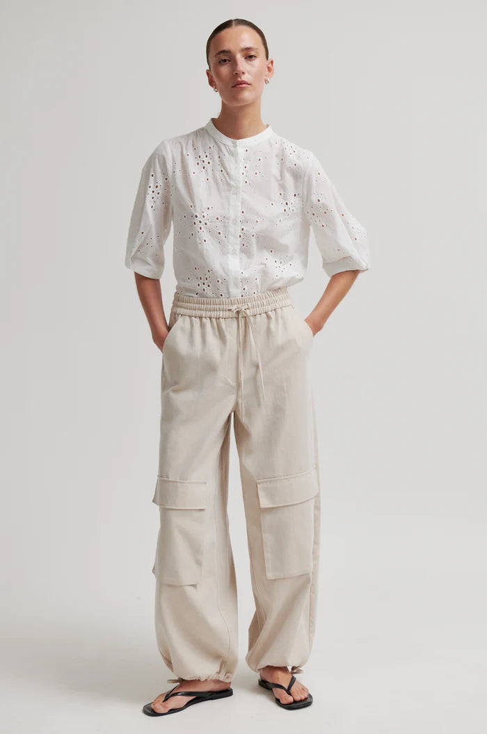 Woman wearing a white blouse with intricate patterns and beige cargo pants on a plain background