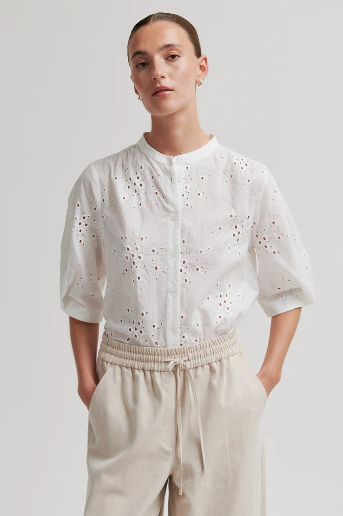 Woman shown up close wearing a white blouse with intricate detailing and beige pants on a light grey background