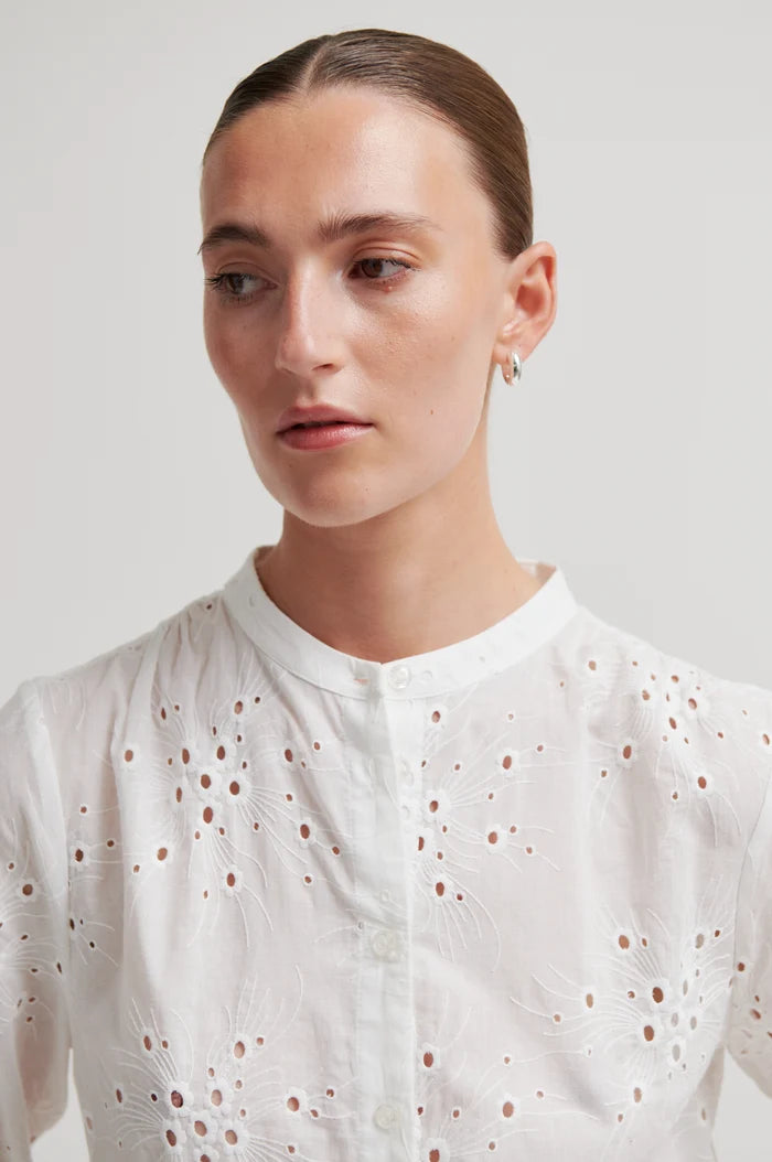 Woman shown up close wearing a white short sleeve blouse with intricate patterns on a plain background