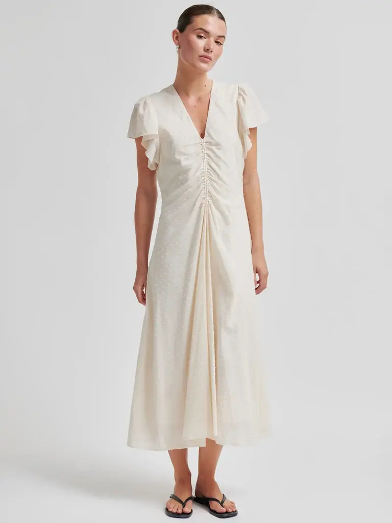 A woman modeling a cream-colored midi dress with short flutter sleeves, a V-neckline, and a gathered front detail, paired with black flip-flop sandals against a light gray studio background.