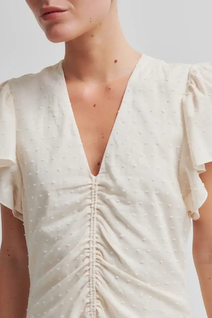Close-up of a model wearing a cream-coloured short-sleeve dress with a deep V-neckline, gathered ruching down the centre front, and a textured dotted fabric.