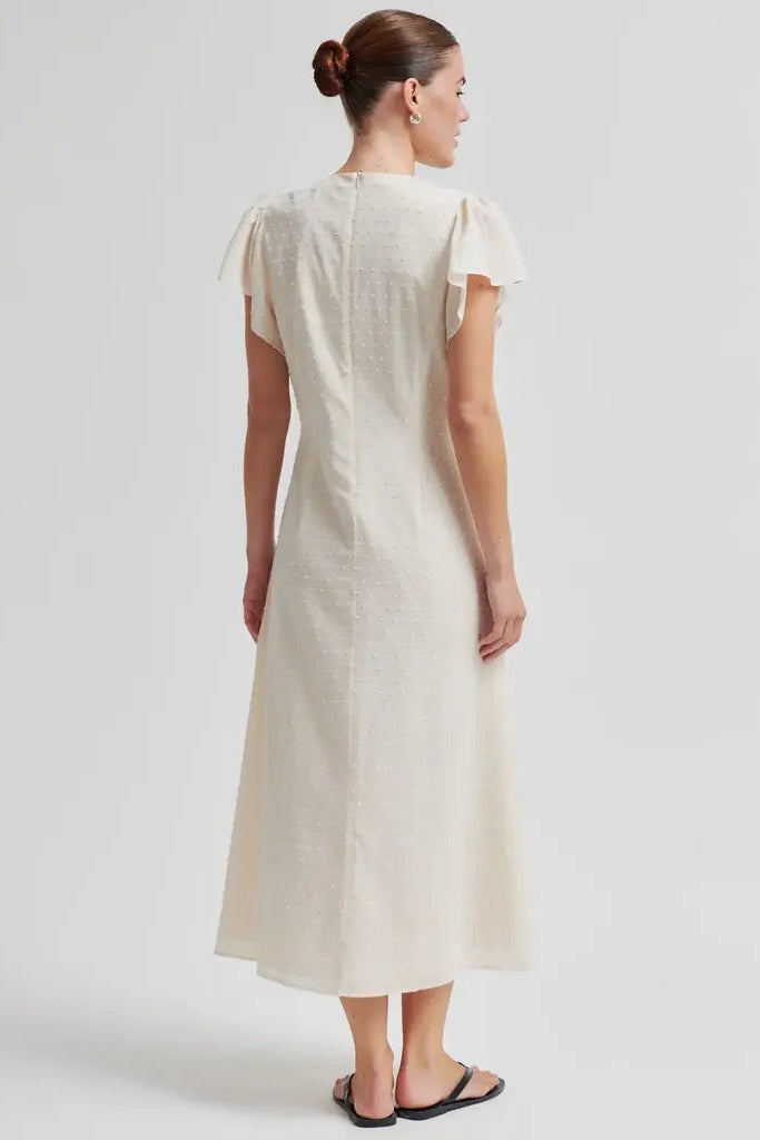 A woman viewed from the back wearing a cream-colored midi dress with ruffled short sleeves and a textured fabric, paired with black flat sandals against a light gray studio background.