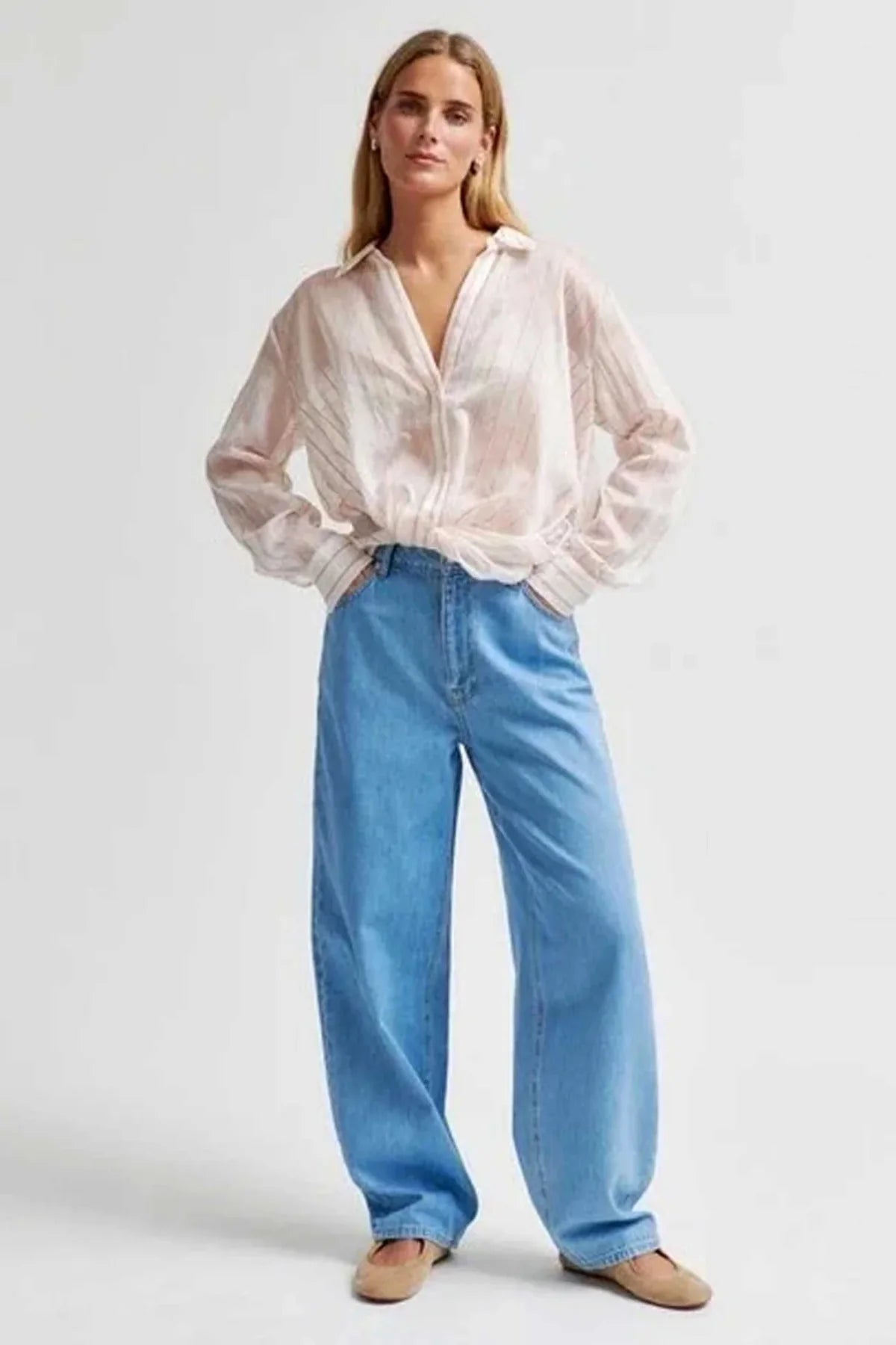 A woman modelling a light pink sheer long-sleeve button-up blouse with twist tie detail and wide-leg light blue jeans, paired with tan flats, against a plain light grey studio background.