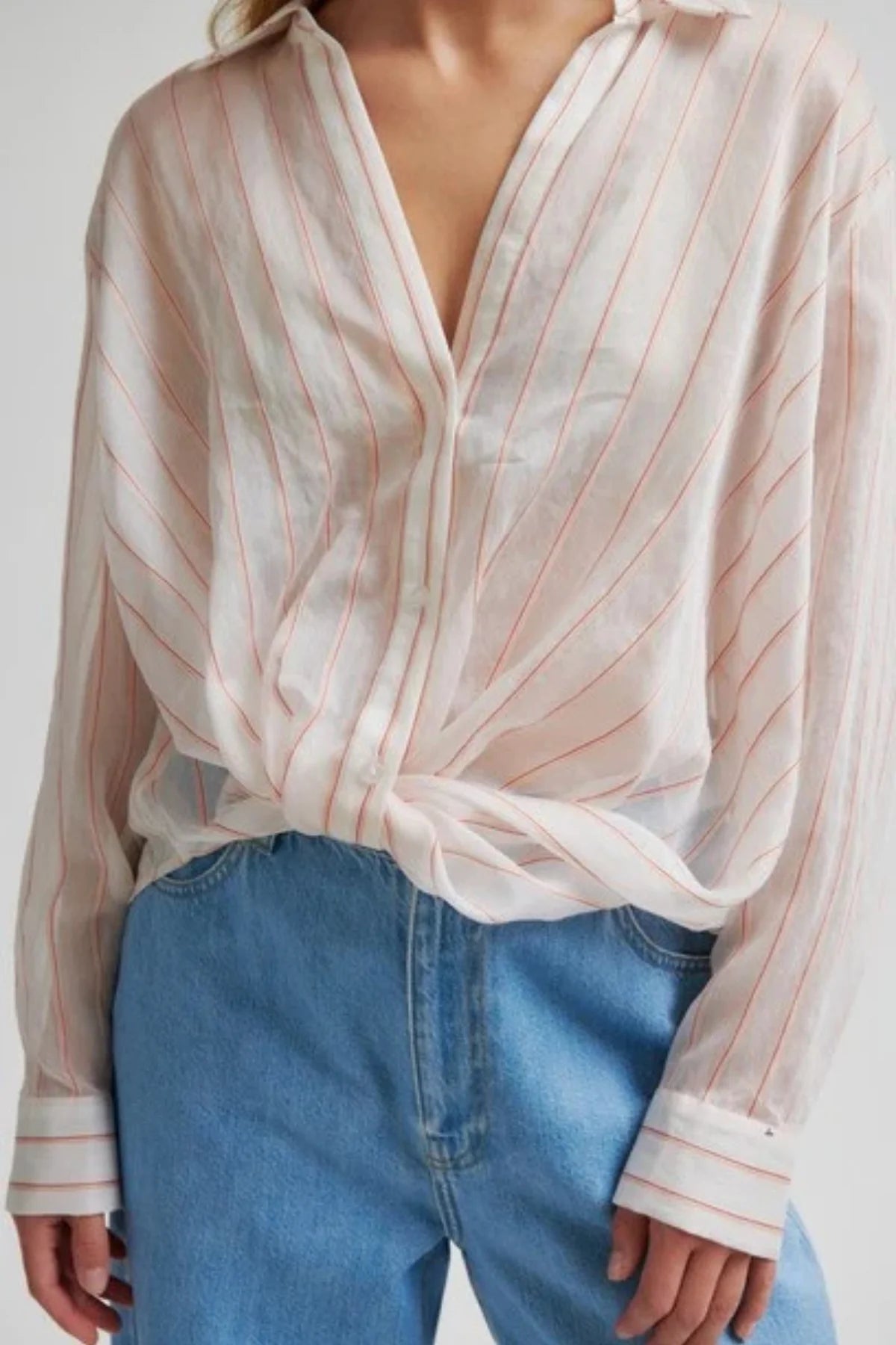 Woman up close wearing a sheer striped blouse with twist front detail and blue jeans against a white background